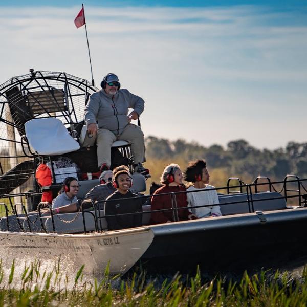 A lively airboat glides through lush marshes at Boggy Creek Adventures in Kissimmee, Florida, with a guide and guests wearing ear protection, enjoying a thrilling Everglades exploration.