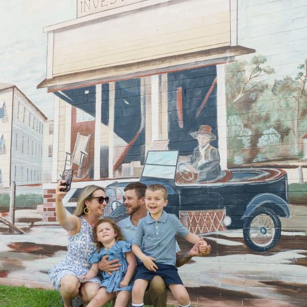 A smiling family poses for a selfie in front of a vintage-style mural in Downtown St. Cloud, Florida, depicting a historic scene with a woman in a car and the old Hotel St. Cloud.