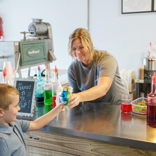 A smiling employee at Sweet Spot Snoballs in Downtown St. Cloud hands a colorful rainbow snoball to a young boy at the counter, surrounded by bottles of flavored syrups.