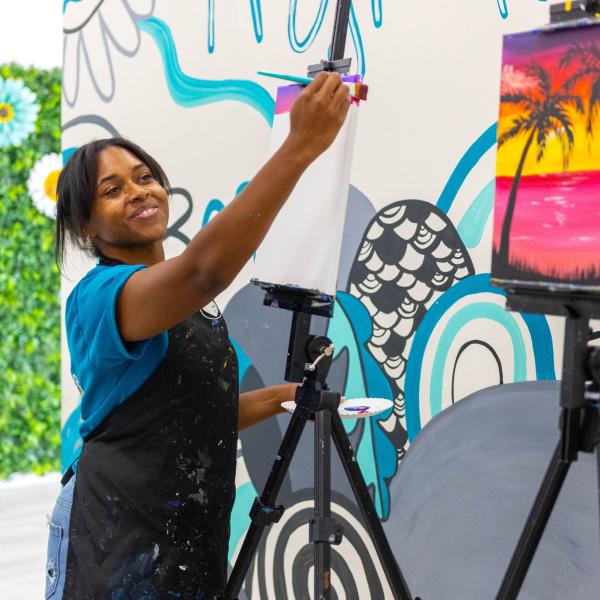 Woman smiling while painting a sunset-themed canvas on an easel at Ex Animo Creations in St. Cloud, Florida.