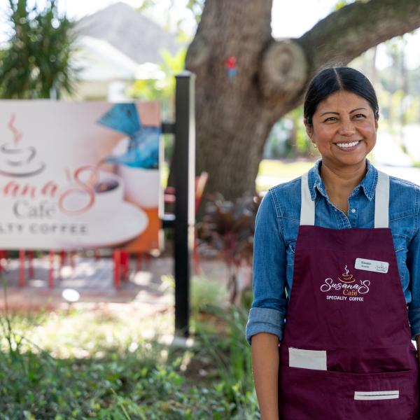 Susana, owner of Susana’s Café, stands smiling in front of her café’s sign and a large oak tree, wearing a burgundy apron that reads “Susana’s Café Specialty Coffee.”