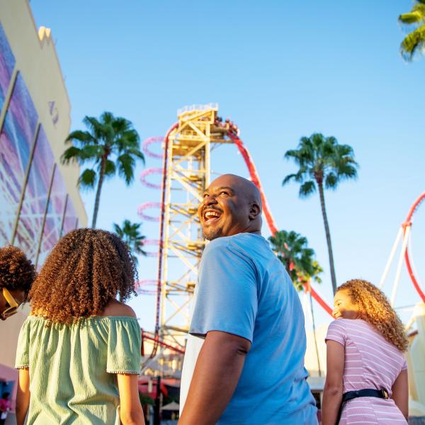 Family smiling and walking toward a towering red roller coaster under a bright blue sky at a theme park in Kissimmee, Florida.