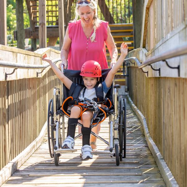 Child using a wheelchair with a zipline harness raises their arms in excitement while being assisted down an accessible wooden ramp at Gatorland in Kissimmee, Florida.