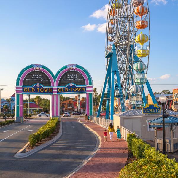 Entrance to Old Town Kissimmee, Florida, featuring the colorful archway and Ferris wheel against a bright blue sky, with visitors walking along the brick pathway.