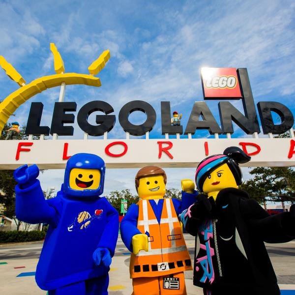 LEGO movie characters pose in front of the colorful entrance sign at LEGOLAND® Florida Resort on a bright, sunny day.