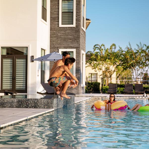 A family enjoys a sunny afternoon at a Kissimmee vacation home pool, with a father and son jumping in while two women relax on inflatable floats.