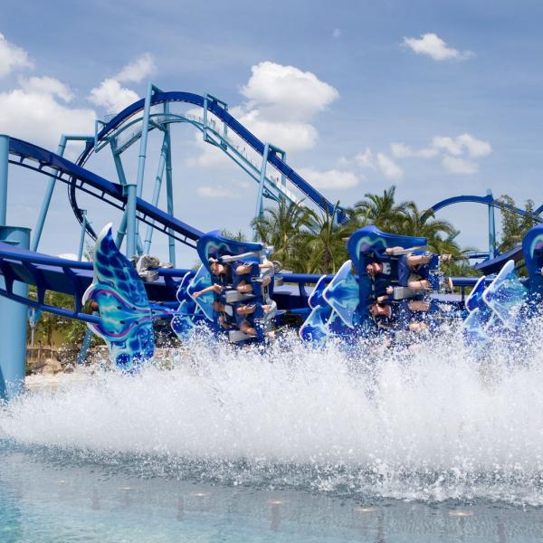 Riders soar over water on SeaWorld Orlando’s Mako-themed coaster as it dips and splashes, creating a dramatic spray beneath the bright blue track.