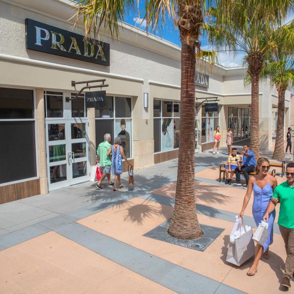 Shoppers stroll along a palm-lined outdoor shopping promenade, passing luxury storefronts while carrying branded shopping bags on a sunny day.
