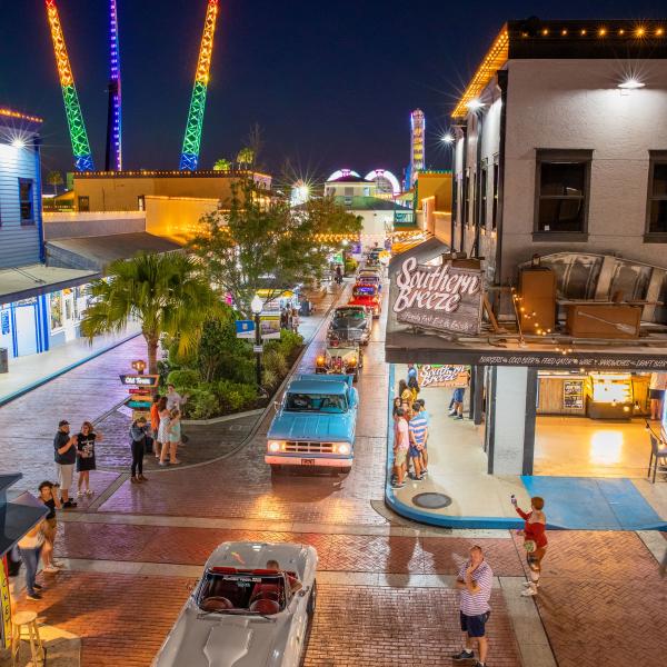A nighttime classic car cruise through Old Town Kissimmee, with colorful neon lights, people watching from the sidewalks, and illuminated thrill rides glowing in the background.