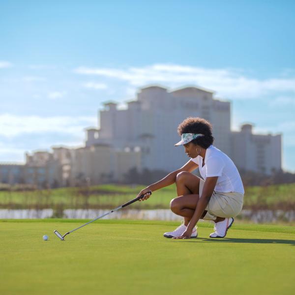 A woman kneels on the putting green of a golf course, aligning her shot with a putter as a large resort building stands blurred in the background.
