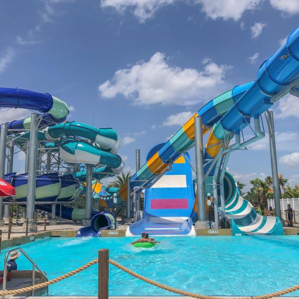Colorful water slides twist and turn above a bright blue splash pool as a rider on a green tube glides into the water at an outdoor water park.