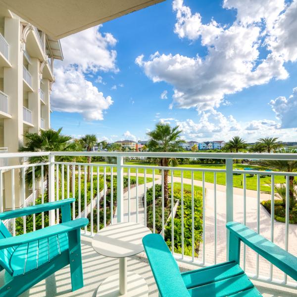 A private balcony with turquoise Adirondack chairs overlooking a sunny resort landscape with palm trees, walking paths, colorful buildings, and a distant lake under a bright blue sky.