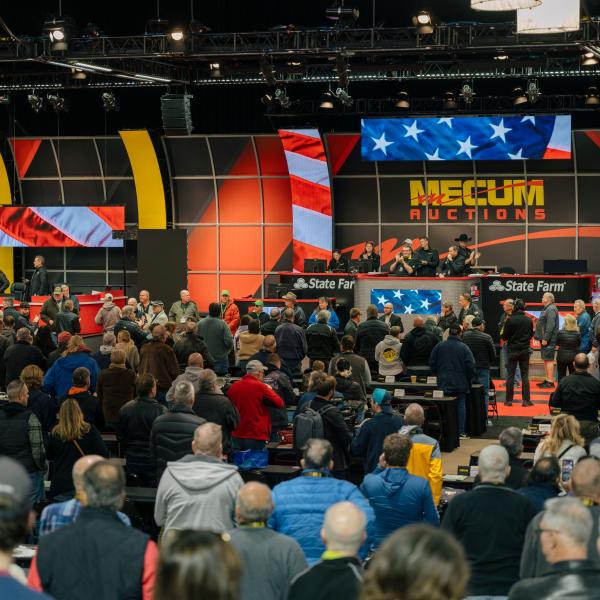 A packed Mecum Auctions hall filled with bidders gathered around the main stage, framed by large American flag displays and bright auction screens.