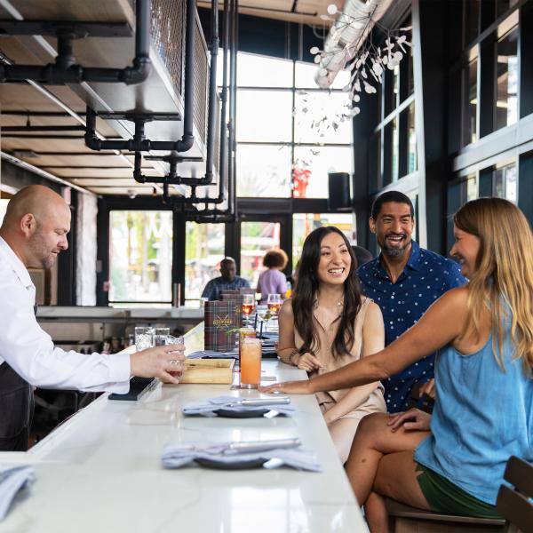 A bartender serves drinks to three smiling guests seated at a modern bar with large windows, creating a bright and lively atmosphere.