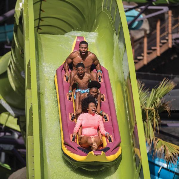 A family of five rides a multi-person raft down a steep green water slide at Universal’s Volcano Bay, splashing through water with excited expressions.