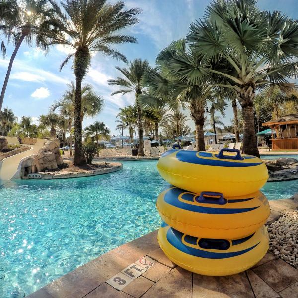A resort-style pool with clear blue water, a rock waterslide, palm trees, and a stack of yellow inflatable tubes beside the pool deck on a sunny day.