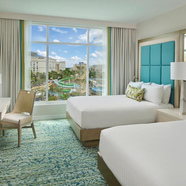 Bright, modern hotel room with two queen beds, teal headboards, a desk, and a large window overlooking the Gaylord Palms water park in Kissimmee, Florida.