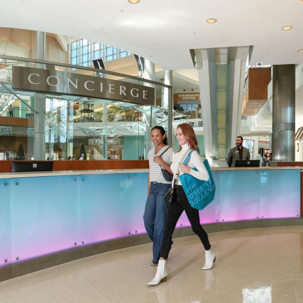 Two women walk past the concierge desk inside The Mall at Millenia, carrying shopping bags in a bright, modern interior with glass accents and high ceilings.