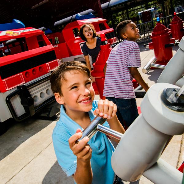 Children play at the Rescue Academy attraction at LEGOLAND® Florida Resort, spraying water from hoses toward LEGO fire trucks in a hands-on firefighting adventure.