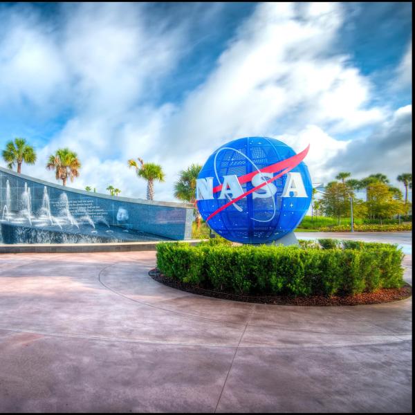 Entrance to Kennedy Space Center Visitor Complex in Merritt Island, Florida, featuring the iconic blue NASA globe and the fountain-lined Space Mirror Memorial under a bright blue sky.