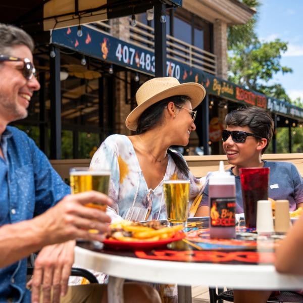 A family enjoying an outdoor meal together at a restaurant patio, smiling and talking over drinks and plates of food on a sunny day.