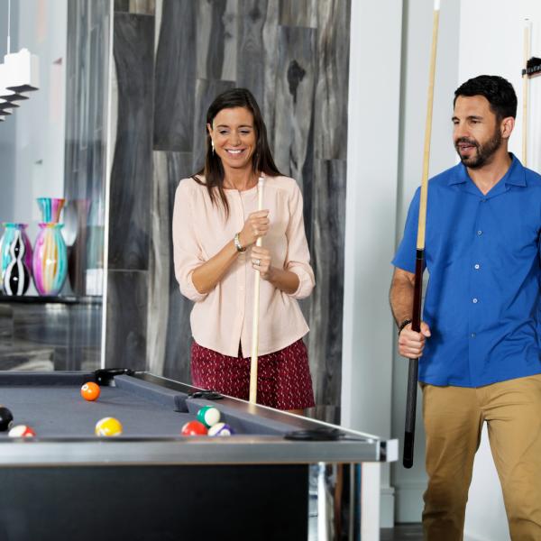 A couple plays a game of pool inside a modern vacation home game room, holding cue sticks beside a billiards table.