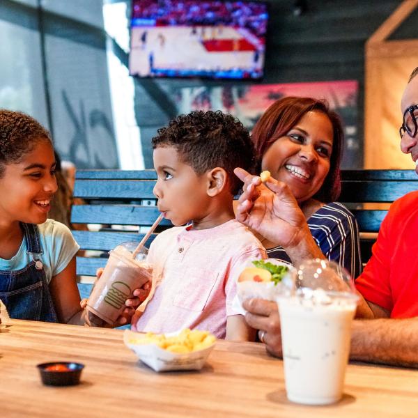Family enjoying a casual meal together at a restaurant, sharing burgers, fries, and milkshakes while smiling and talking around a wooden table.