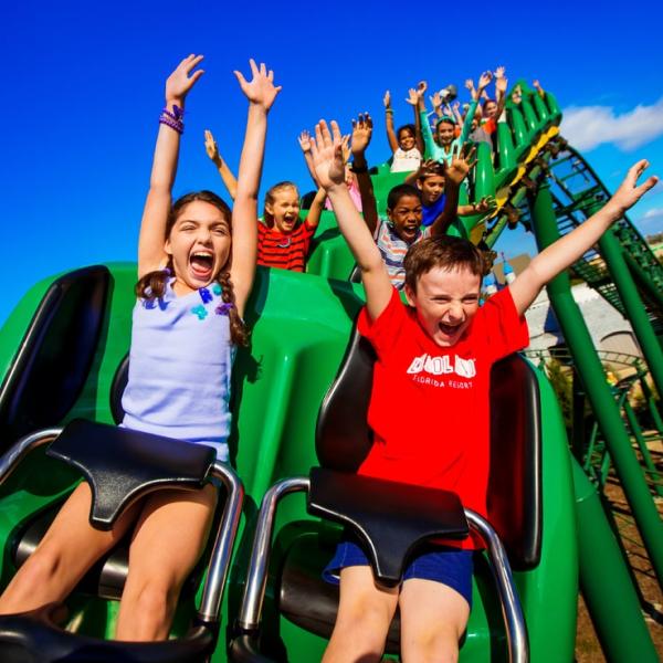 Children ride a bright green roller coaster with their arms raised, laughing and cheering under a clear blue sky.