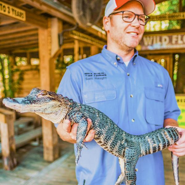 A Wild Florida staff member smiles while holding a small alligator in an outdoor educational exhibit area.