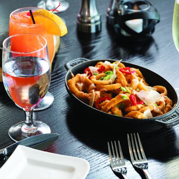 A close-up of a restaurant table setting featuring a dish of fried calamari topped with peppers and herbs, accompanied by drinks including water, a cocktail, and a glass of sparkling wine.