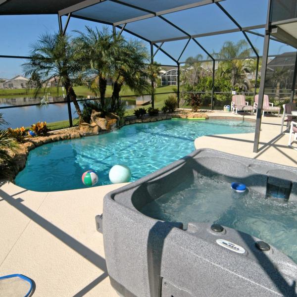 Screened-in private pool and hot tub at a Kissimmee vacation home, featuring tropical landscaping, outdoor dining seating, and a peaceful pond view.