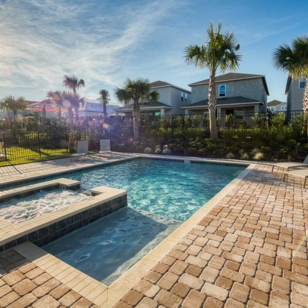 Private vacation home pool with a built-in spa, stone paver deck, lounge chairs, and palm trees, set against a quiet residential backdrop at sunset.