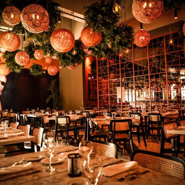 A warmly lit room with banquet seating, mirror accents, and round wicker lighting and ferns adorning the ceiling.