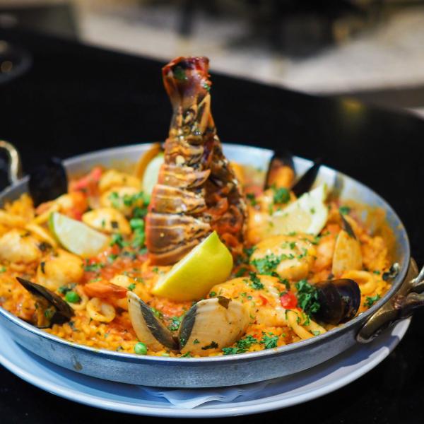 A pan of seafood paella topped with a lobster tail, mussels, shrimp, scallops, lemon wedges, and fresh herbs sits on a white plate on a dark tabletop.