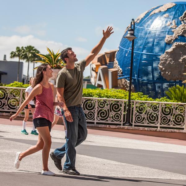 Couple walks through Universal Orlando Resort, smiling and waving near the iconic globe entrance.
