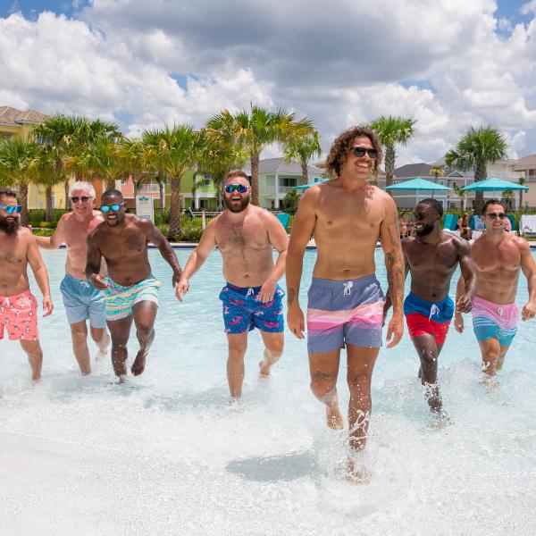 A group of men walks through the shallow end of a resort pool on a sunny day, smiling and enjoying their time together.
