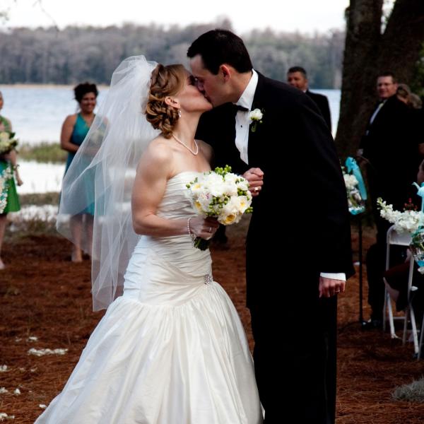 A bride and groom share a kiss at their outdoor wedding ceremony beside a lakeshore, surrounded by guests and bridesmaids.