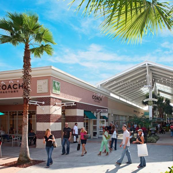 Shoppers stroll through an outdoor shopping plaza lined with palm trees and brand-name stores at Orlando Premium Outlets near Kissimmee, Florida.