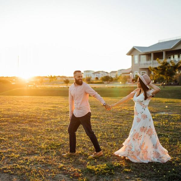 Couple holding hands and walking through a sunlit field at Island Grove Wine Company in Kissimmee, Florida, with golden light and vineyard-style buildings in the background.
