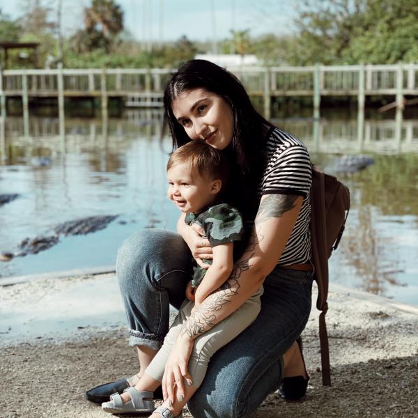 @IzzyRose kneels beside her young child near the water at Gatorland near Kissimmee, Florida, smiling as they enjoy a sunny day surrounded by nature and wildlife.