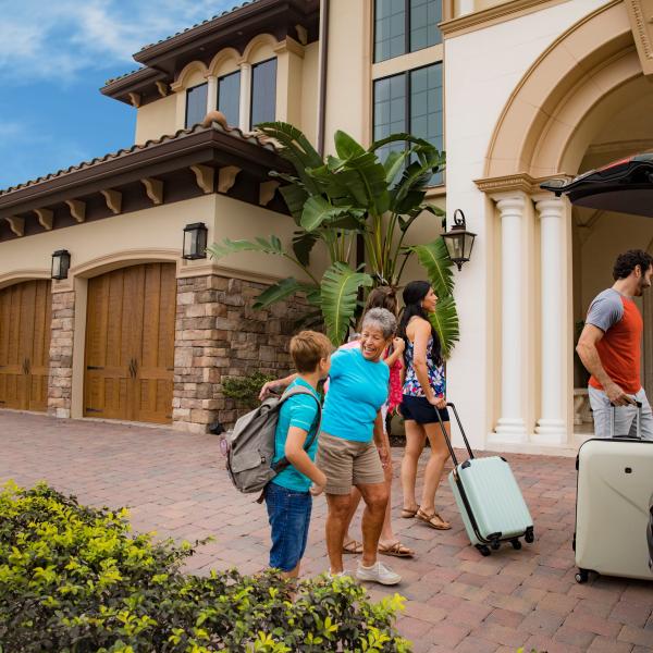 A multi-generational family arrives at a luxury vacation home in Kissimmee, Florida, smiling as they unpack luggage from their car on a sunny day.