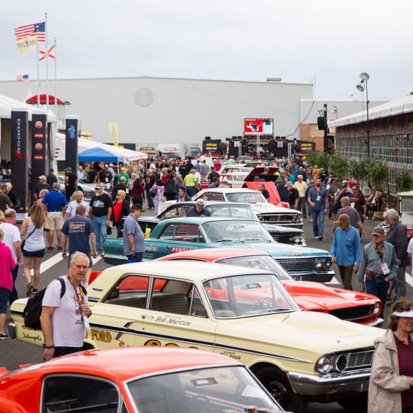 A large crowd strolls through rows of classic and vintage cars at a busy outdoor auto auction, with colorful muscle cars on display and vendor tents lining the walkway.