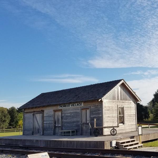 Historic wooden buildings at Pioneer Village sit under a bright blue sky, surrounded by grassy fields, trees, and a gravel walkway.