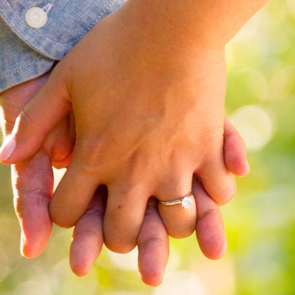 Two people hold hands outdoors, with a diamond engagement ring visible against a softly blurred green background.