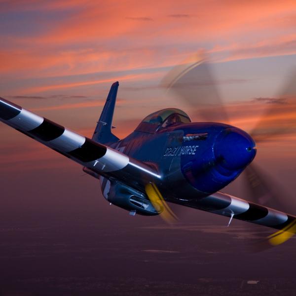 Blue P-51 Mustang aircraft named “Crazy Horse” soars through the sky at sunset, its propeller in motion against a vivid orange and purple horizon.