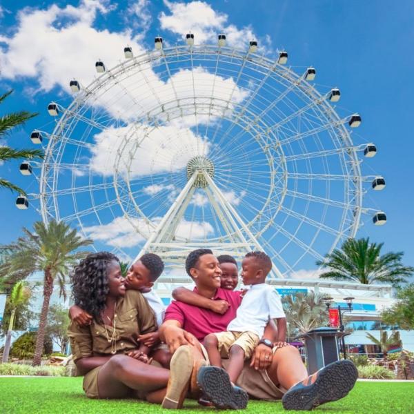 A family of five sits together on the grass in front of The Wheel at ICON Park, smiling and embracing on a bright, sunny day.