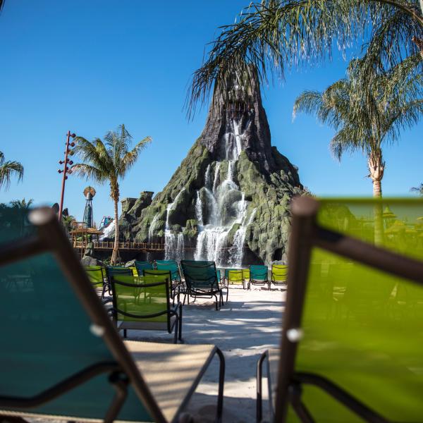 Lounge chairs on a sandy beach face Krakatau Volcano with cascading waterfalls at Universal’s Volcano Bay, framed by palm trees under a clear blue sky.