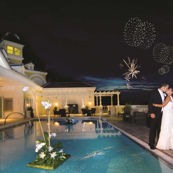A bride and groom kissing beside a pool and house at night with fireworks going off at night.