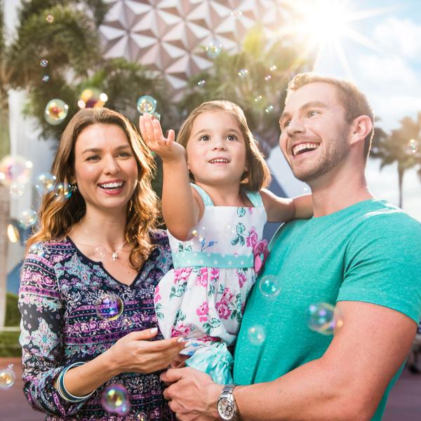 A couple holds their child in their arms as they visit Epcot Center and she reaches for bubbles passing.