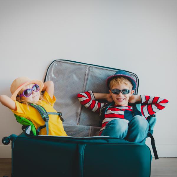 Two young children wearing sunglasses and travel outfits lounge playfully inside an open suitcase, pretending they’re ready for vacation.